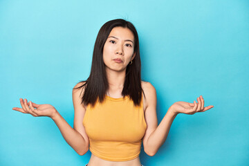 Asian woman in summer yellow top, studio setup, doubting and shrugging shoulders in questioning...