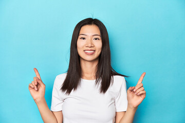 Young Asian woman in white t-shirt, studio shot, indicates with both fore fingers up showing a...