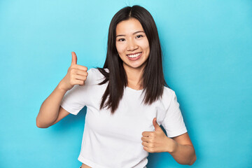 Fototapeta premium Young Asian woman in white t-shirt, studio shot, raising both thumbs up, smiling and confident.