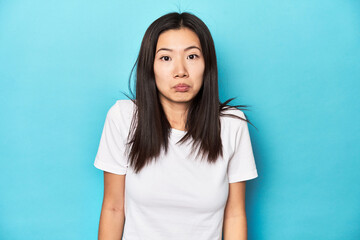 Young Asian woman in white t-shirt, studio shot, shrugs shoulders and open eyes confused.