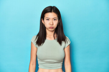 Asian woman in summer green top, studio backdrop, blows cheeks, has tired expression. Facial...