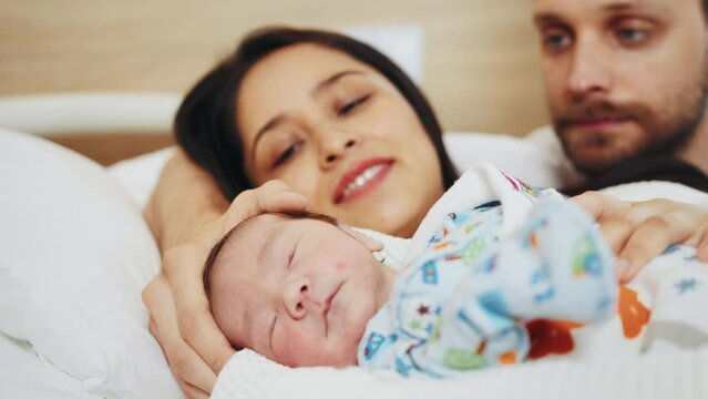 Newborn Baby Boy Being Cradled By New Mother And Father In Birthing Center. Joy And Happiness Emotions At Faces Of New Parents. Smiling Mother Sitting In Hospital Bed After Childbirth.