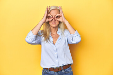 Middle-aged Caucasian woman in blue shirt, yellow studio showing okay sign over eyes