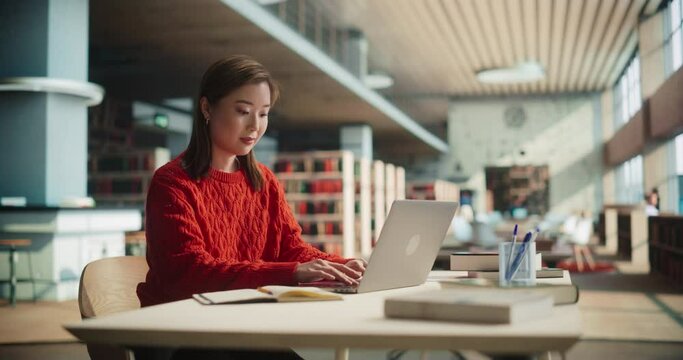 Portrait Shot of an Asian Female in a Red Coat Creating a Project Plan for a Class Assignment, Writing in Notebook and Using a Laptop Computer in a Modern Public Library Reading Hall