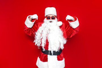 strong santa claus in red glasses shows biceps on colored background, man in hat and santa costume for christmas