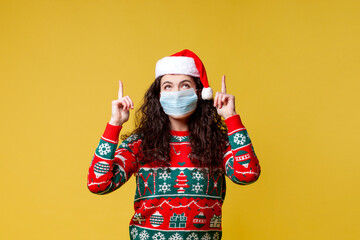 Fototapeta na wymiar young girl in christmas clothes and santa hat in medical protective mask shows her hands up on yellow background