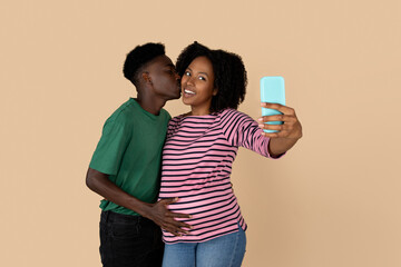 Naklejka premium Positive millennial black guy touching lady belly, kiss, enjoy pregnancy, take selfie on smartphone