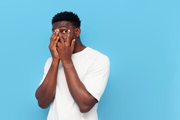 frightened young guy african american in white t-shirt covers his face with his hands and peeps on blue background
