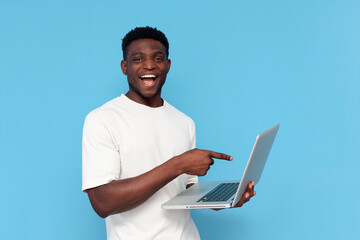 african guy in white t-shirt uses laptop and smiles on blue isolated background, man points his finger at the screen