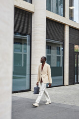 Young AFrican American businessman in white suit moving down sidewalk in urban environment along modern architecture with large windows