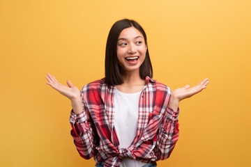 Cheerful japanese lady shrugging shoulders, looking aside and smiling, standing over yellow studio background