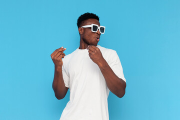 african american man in white t-shirt and sunglasses dances to music and sings on blue isolated...