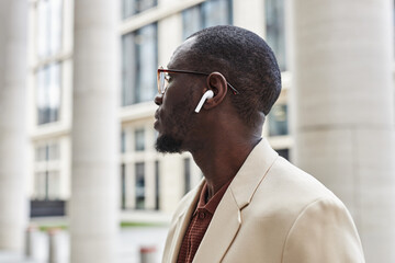 Side view of young serene black man in elegant white suit, eyeglasses and earphones listening to...