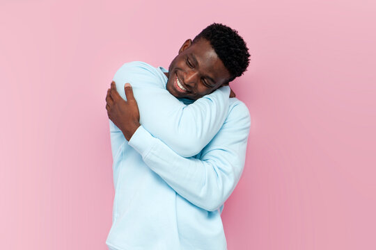 african american man in blue warm sweater hugs and loves himself on pink isolated background