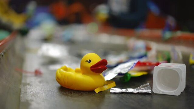 Close-up yellow rubber duck on conveyor belt with garbage and blurred workers sorting trash at background in slow motion. Toy and litter on factory for recycling indoors