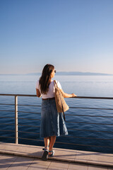 Back view of attractive woman in denim style stand in front of sea landscape. Happy woman looking on sunset