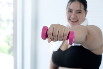 Happy overweight  woman doing exercise with dumbbells over white wall at home, wearing sports wear, looking at camera, selective focus