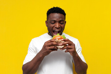 african american man in white t-shirt bites big burger on yellow isolated background, young guy...