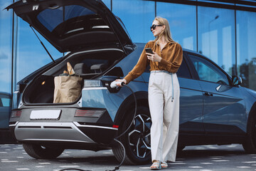 Woman with food shopping bags charging electric car and using mobile phone