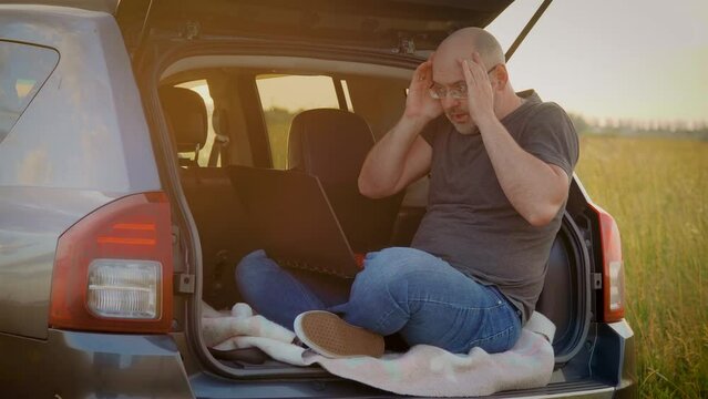 Guy with laptop types on computer and commits irreparable mistake in commercial project, man outside works remotely, sitting in car trunk while traveling. Busy worker freelancer entrepreneur notebook