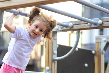 Fototapeta premium child sport. little active girl on playground in park, hanging on horizontal bar, rope ladder, workout, doing sports exercise outdoors. happy childhood. Kid play on school kindergarten yard in summer