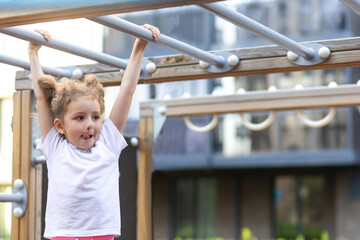 child sport. little active girl on playground in park, hanging on horizontal bar, rope ladder, workout, doing sports exercise outdoors. happy childhood. Kid play on school kindergarten yard in summer