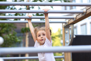 Fototapeta premium child sport. little active girl on playground in park, hanging on horizontal bar, rope ladder, workout, doing sports exercise outdoors. happy childhood. Kid play on school kindergarten yard in summer