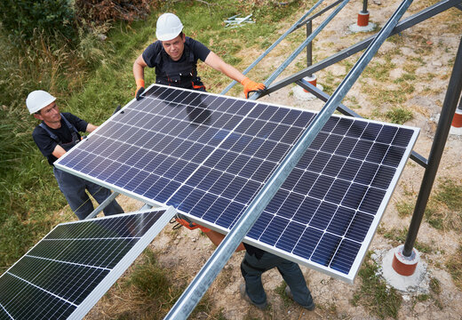 Workers mounting photovoltaic solar panel system outdoors. Men engineers placing solar module on metal rails, wearing construction helmets and work gloves. Renewable and ecological energy.