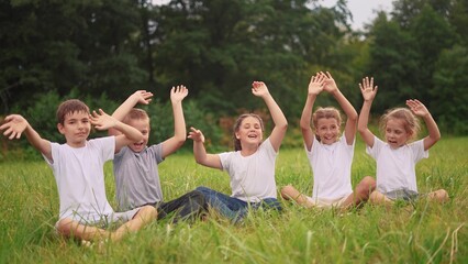 Naklejka premium children sit on the grass in the park with their hands up. happy family childhood dream concept. kids are resting in a park in the meadow sitting on the lifestyle grass and stretching their hands up