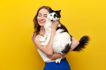 young cute girl holding black and white cat on yellow isolated background and smiling, woman with pet