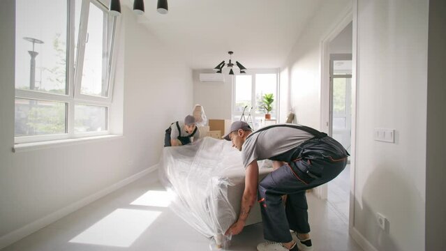 Porter men arranging modern furniture in updated flat. Repairmen establishing couch at apartment. Two men wearing overalls carrying sofa. Industry men finishing renovation at new building.