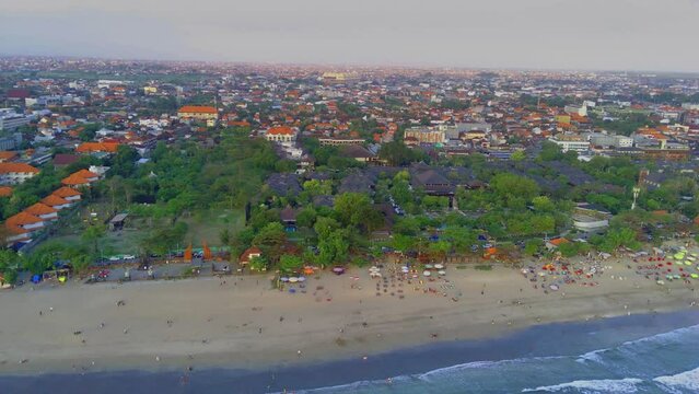 The bustling atmosphere of Legian Beach, Kuta, Bali, with the view of the trees and hotels during a crowded day, captured from the air using a drone. Aerial Video.