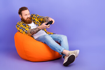 Photo of funky guy man wear trendy clothes sit cozy pouf spend free time playstation watch empty space isolated on purple color background © deagreez