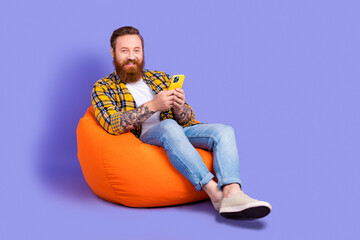 Photo of smiling handsome man wear checkered stylish clothes sitting pouf use modern phone empty space isolated on purple color background