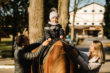 Kid riding a horse. Family having fun spring vacation on farm ranch. Little boy ride in saddle on horse. Happy father, mother, son caress horse outdoors. Dad, mom, child cuddling animal pet at sunset.