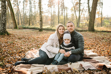 Autumn mood. Mother, father, kid have picnic in autumn forest in nature. Mom, dad hugs son child sitting on blanket on yellow leaves in park. Family spending time together at sunset. Autumn holiday.