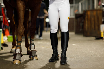 Female legs in black leather boots close up rider jockey walking with horse at stable and preparing horse racing or jumping competition