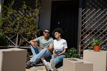 happy african american couple holding coffee and sitting near packages and new house