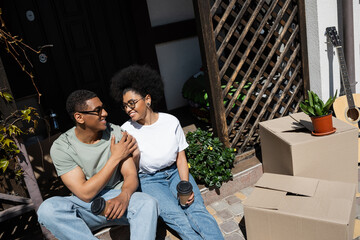 cheerful african american couple with coffee holding hands near carton boxes and new house