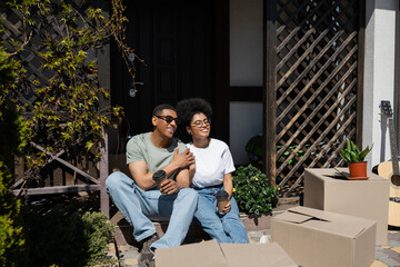 smiling african american couple with coffee talking near carton boxes on porch of new house