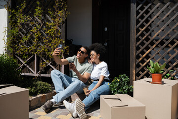 smiling african american couple with coffee to go taking selfie near boxes and new house