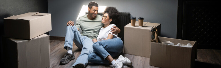 smiling african american couple hugging near carton boxes and takeaway coffee in new house, banner