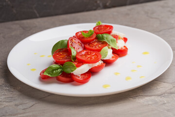 A view of caprese salad with tomatoes, mozzarella, basil and olive oil on white plate on dark stone table. Isometric style. Mediterranean cuisine. Selective focus.