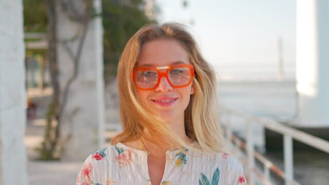 Attractive young woman walking down street near sea confidently looking at camera. Cheerful blonde in floral dress and orange glasses smiling enjoying walk during vacation