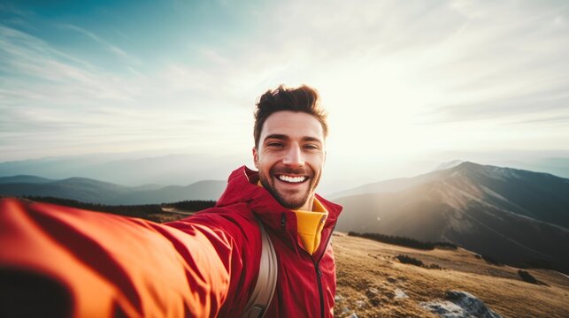a young male hiker with a smile on his face, looking at the camera and capturing a selfie portrait at the mountain summit