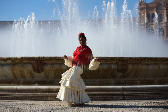 Young black woman dressed as a flamenco gypsy in a famous square in Seville, Spain. She is wearing a beige dress with ruffles and a red shawl and is standing in front of a fountain.