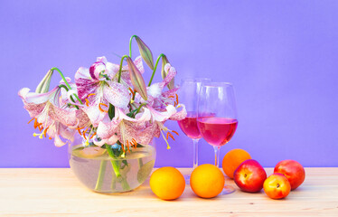 Bouquet of white lilies with purple spots in glass round vase, glasses of rose wine and fresh fruits on wooden table. Purple background.
