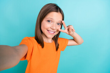 Photo of adorable cheerful little girl wear orange stylish t-shirt doing selfie show v-sign on eye isolated on teal color background