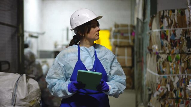 Dolly shot concentrated young woman in uniform and hard hat with digital tablet walking on recycling station leaving. Front view portrait of confident Caucasian worker checking garbage sorting indoors