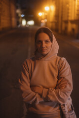 young thoughtful woman stands on road in beige hoodie. walk in evening in light of lanterns, portrait of female alone. freedom loneliness of modern generation. Travel and rest alone, mental health